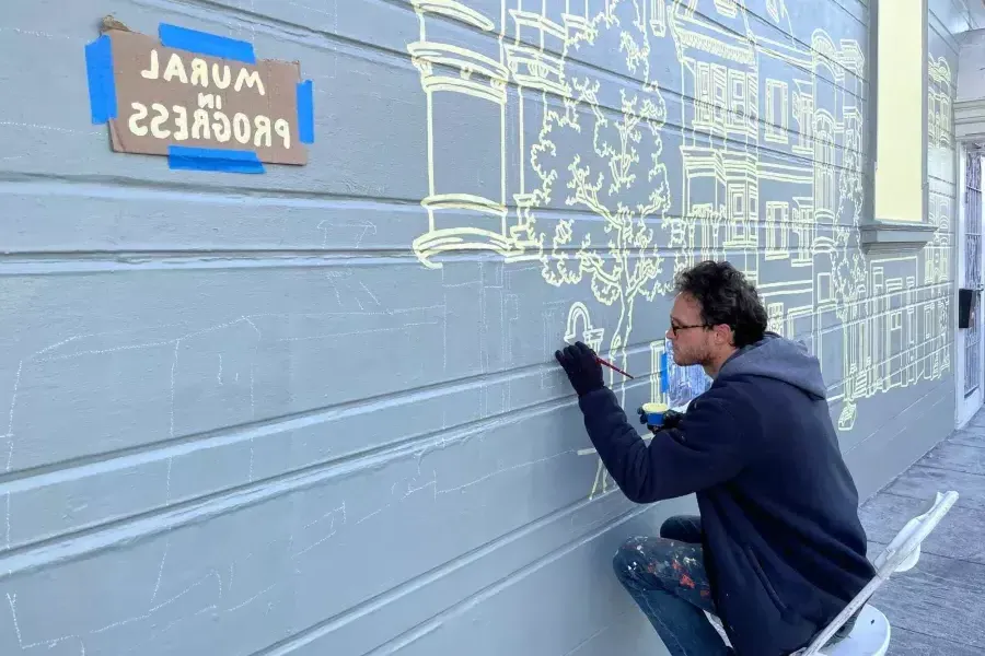 An artist paints a mural on the side of a building in the Mission District, with a sign taped onto the building that reads "Mural in Progress.“是威尼斯人官网平台app,ca.